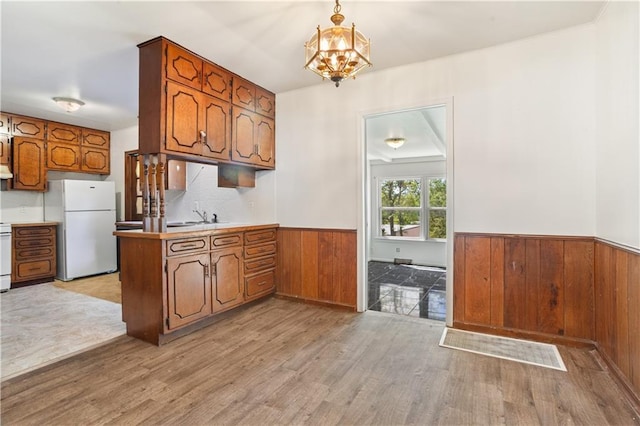 kitchen with a peninsula, freestanding refrigerator, a wainscoted wall, brown cabinetry, and light wood-type flooring