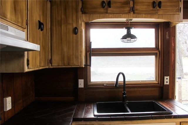 kitchen featuring a sink, brown cabinets, tile countertops, and under cabinet range hood