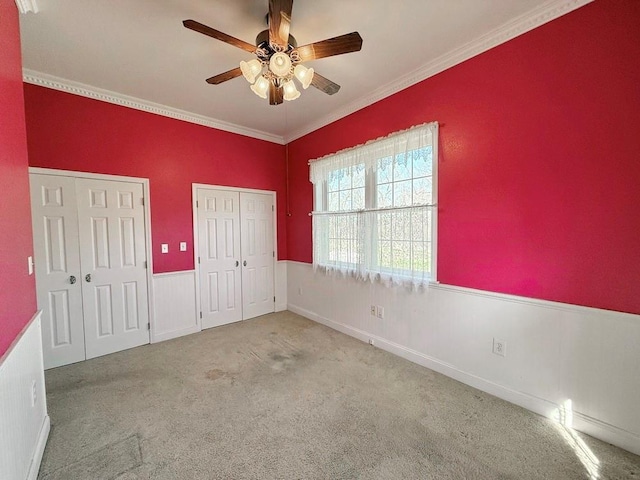 unfurnished bedroom with a ceiling fan, carpet, crown molding, multiple closets, and a wainscoted wall