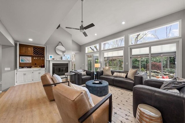 living area with high vaulted ceiling, a glass covered fireplace, light wood-style floors, ceiling fan, and recessed lighting