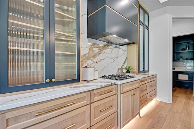 kitchen featuring light brown cabinetry, wall chimney range hood, stainless steel gas stovetop, light stone counters, and light wood finished floors