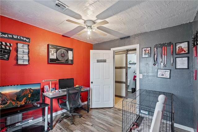 office area with a ceiling fan, wood finished floors, baseboards, visible vents, and a textured wall