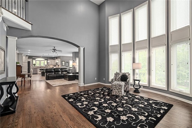 living room with arched walkways, a towering ceiling, decorative columns, a ceiling fan, and wood finished floors