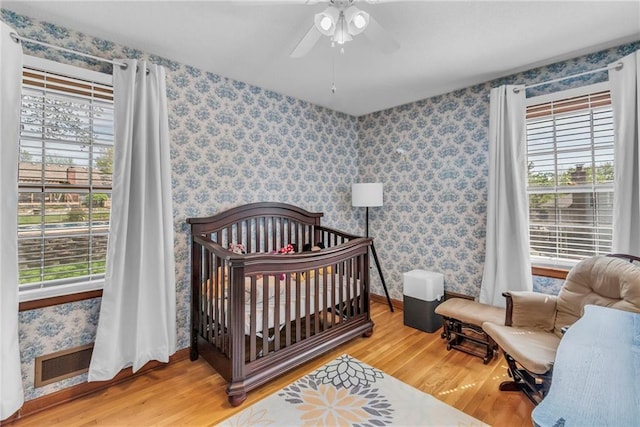 bedroom with visible vents, a nursery area, wood finished floors, baseboards, and wallpapered walls