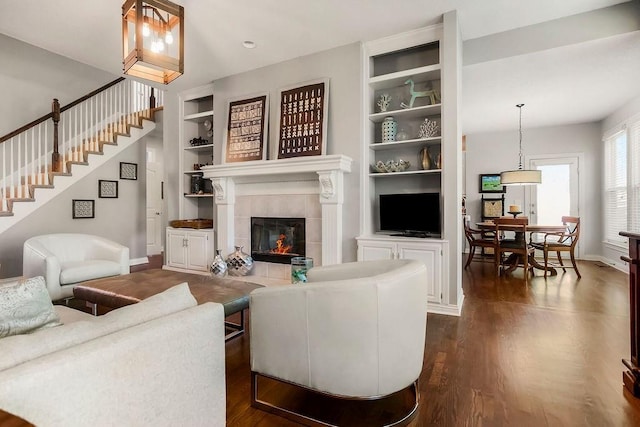 living area featuring dark wood finished floors, baseboards, a tile fireplace, built in shelves, and stairway