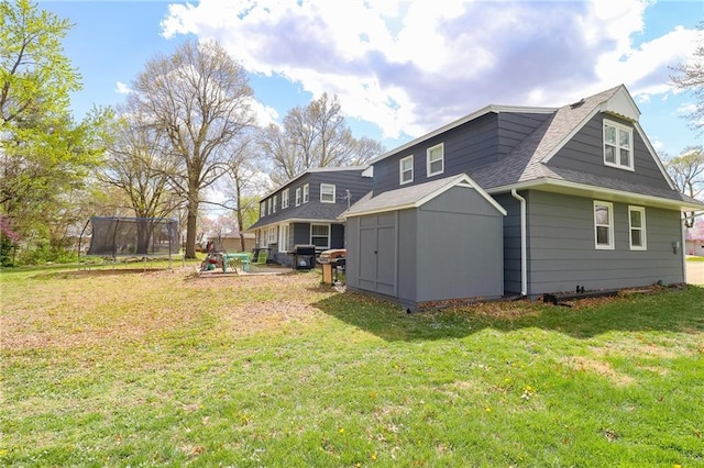 back of property with a trampoline, a lawn, a storage unit, an outbuilding, and roof with shingles