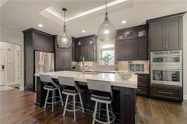 kitchen with appliances with stainless steel finishes, a raised ceiling, a kitchen island with sink, arched walkways, and backsplash