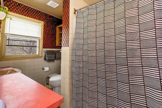 bathroom with a sink, toilet, tile walls, visible vents, and wallpapered walls
