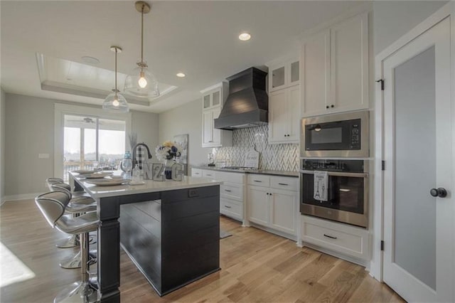 2303 SW Serena Pl, Lees Summit, MO 64082 - kitchen with appliances with stainless steel finishes, custom range hood, a tray ceiling, tasteful backsplash, and light wood finished floors