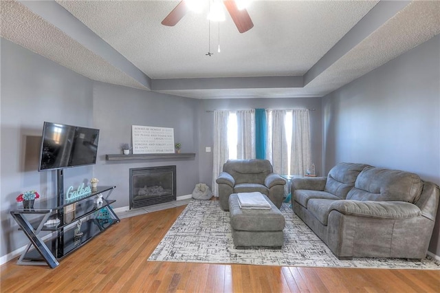 living room featuring wood-type flooring, baseboards, ceiling fan, a tray ceiling, and a fireplace with flush hearth