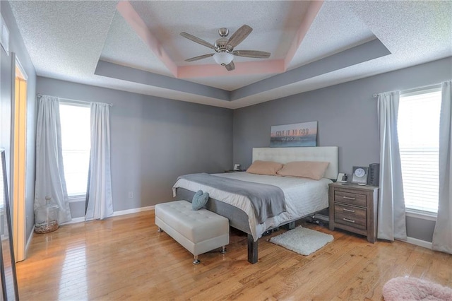 bedroom with baseboards, ceiling fan, a textured ceiling, a tray ceiling, and light wood finished floors