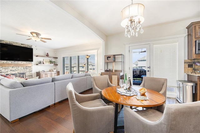 dining area with ceiling fan with notable chandelier, a stone fireplace, and dark wood-type flooring