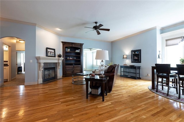 living area with ornamental molding, arched walkways, light wood-type flooring, a glass covered fireplace, and ceiling fan