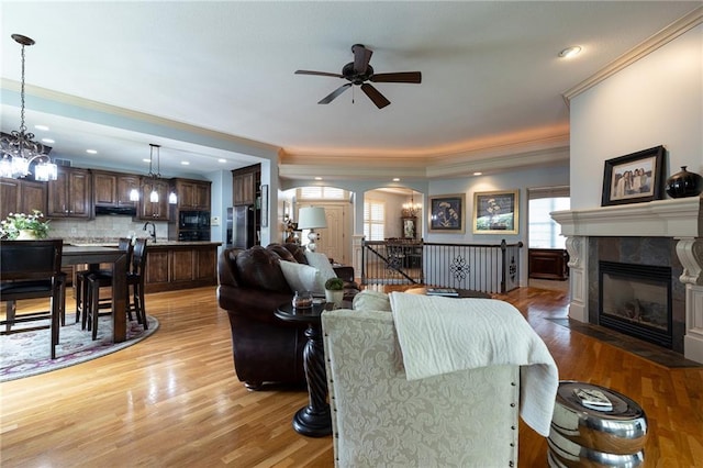 living room with light wood finished floors, arched walkways, a tiled fireplace, ceiling fan with notable chandelier, and crown molding
