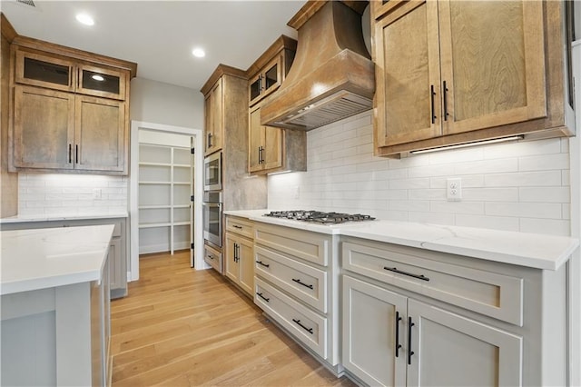 kitchen featuring custom range hood, stainless steel appliances, light wood-type flooring, glass insert cabinets, and decorative backsplash