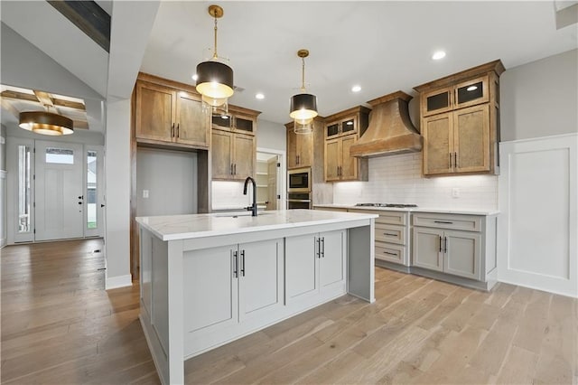 kitchen with premium range hood, appliances with stainless steel finishes, brown cabinetry, a center island with sink, and recessed lighting