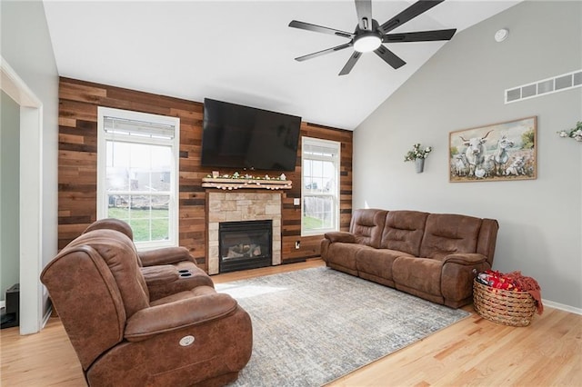 living room with wood walls, high vaulted ceiling, wood finished floors, ceiling fan, and a stone fireplace