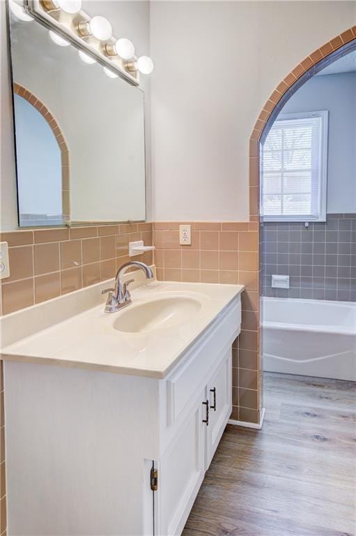 full bath featuring tile walls, a bathtub, vanity, wood finished floors, and a wainscoted wall