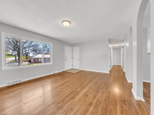 unfurnished living room featuring arched walkways, a textured ceiling, visible vents, light wood finished floors, and baseboards