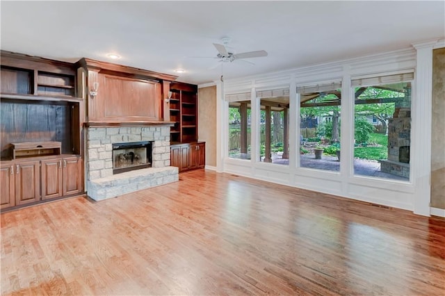 unfurnished living room featuring ceiling fan, a stone fireplace, crown molding, baseboards, and light wood-style floors