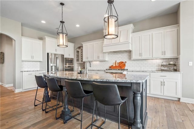 kitchen featuring arched walkways, appliances with stainless steel finishes, an island with sink, and white cabinets