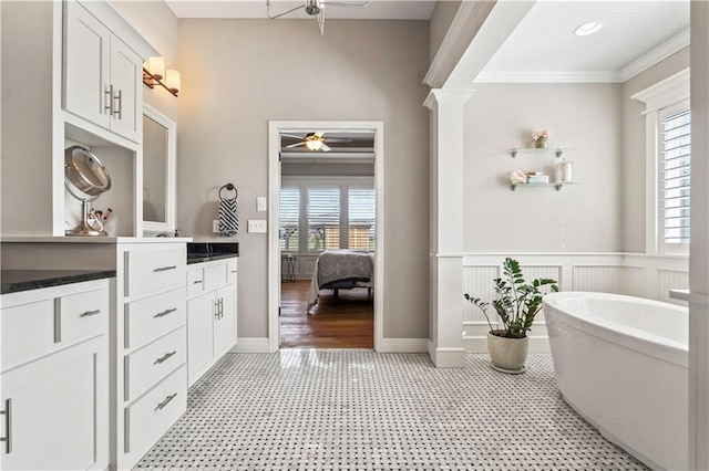 full bath with a soaking tub, crown molding, a ceiling fan, a wainscoted wall, and decorative columns