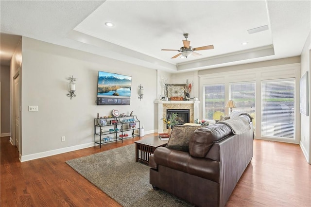 living room featuring a glass covered fireplace, wood finished floors, baseboards, ceiling fan, and a raised ceiling