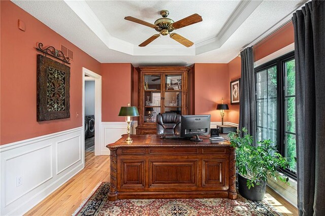 home office featuring washer and dryer, ceiling fan, wainscoting, light wood-style floors, and a tray ceiling