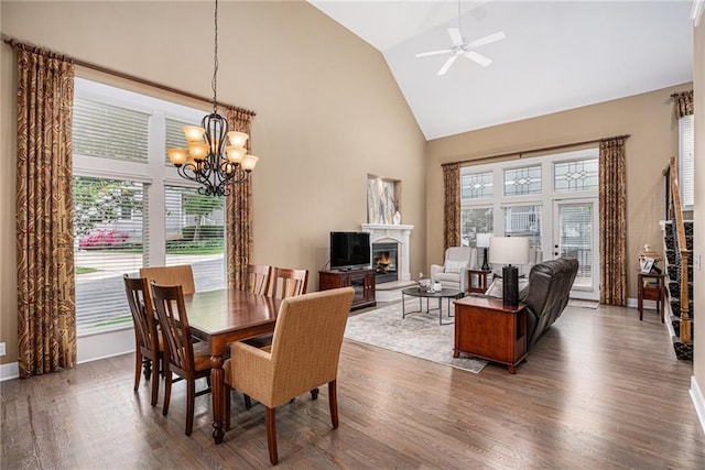 dining space with high vaulted ceiling, a glass covered fireplace, ceiling fan with notable chandelier, baseboards, and wood finished floors