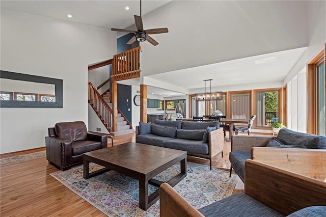 living room featuring light wood-type flooring, stairway, baseboards, ceiling fan with notable chandelier, and a high ceiling