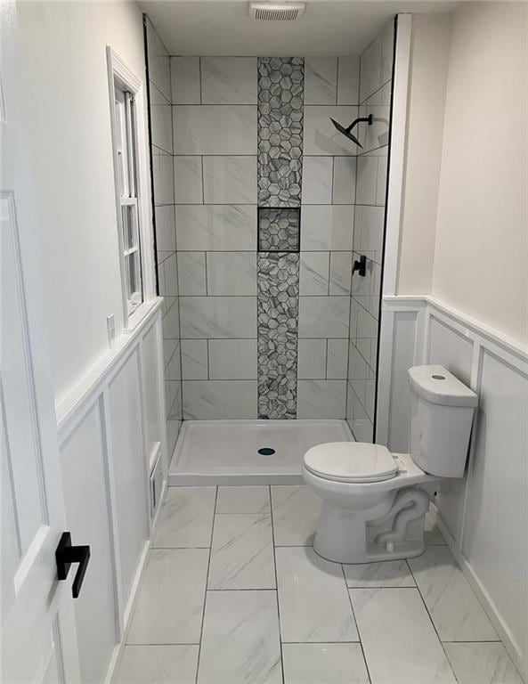 bathroom featuring marble finish floor, wainscoting, a decorative wall, toilet, and tiled shower
