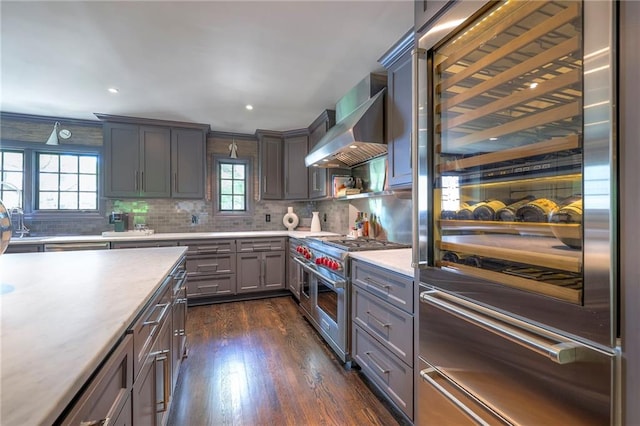 kitchen featuring light countertops, wall chimney exhaust hood, high end appliances, decorative backsplash, and dark wood-style floors