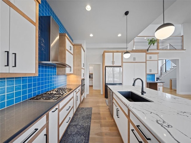 kitchen with hanging light fixtures, appliances with stainless steel finishes, a sink, light wood-style floors, and wall chimney exhaust hood