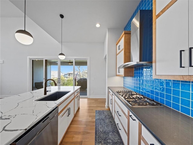 kitchen with light wood-style floors, wall chimney exhaust hood, light stone counters, a sink, and stainless steel appliances