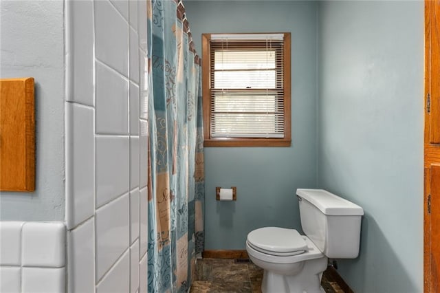 bathroom featuring curtained shower, stone finish flooring, toilet, and baseboards