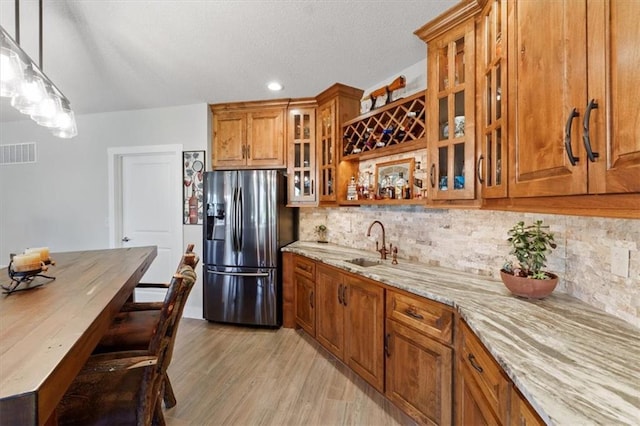 kitchen with refrigerator with ice dispenser, brown cabinetry, light stone counters, backsplash, and a sink
