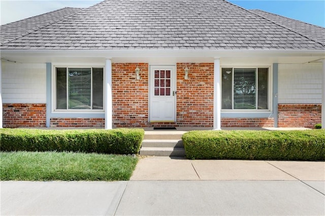 entrance to property featuring brick siding, a shingled roof, and covered porch