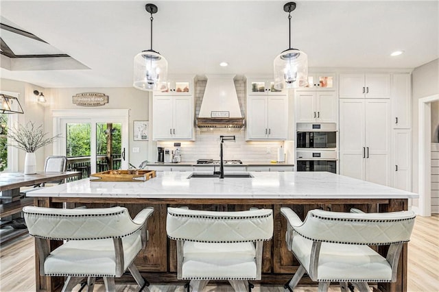 kitchen featuring stainless steel double oven, a center island with sink, light wood finished floors, custom range hood, and white cabinetry