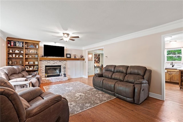 living room with a ceiling fan, crown molding, a brick fireplace, baseboards, and light wood-style flooring