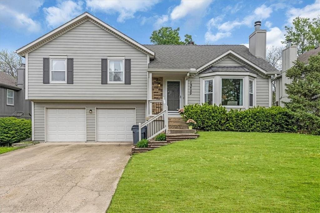 split level home with a chimney, roof with shingles, a front yard, concrete driveway, and an attached garage