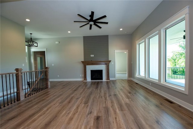 unfurnished living room with baseboards, visible vents, ceiling fan with notable chandelier, a brick fireplace, and wood finished floors