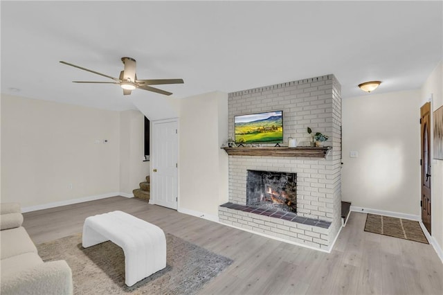 living area with stairs, a brick fireplace, wood finished floors, baseboards, and ceiling fan