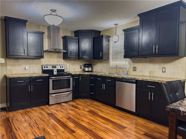 kitchen with light stone counters, wall chimney exhaust hood, stainless steel appliances, a sink, and wood finished floors