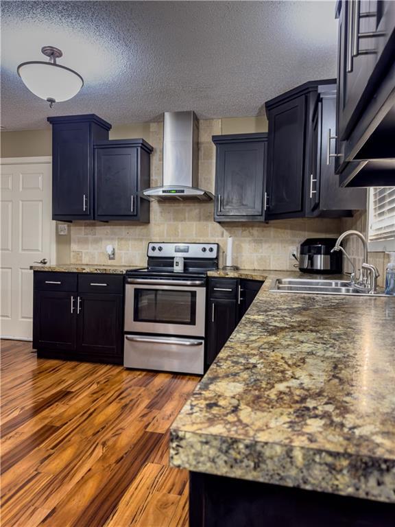 kitchen with stainless steel electric stove, wall chimney exhaust hood, a textured ceiling, a sink, and backsplash