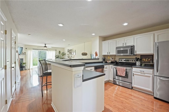 kitchen with light wood finished floors, stainless steel appliances, a peninsula, dark countertops, and white cabinetry