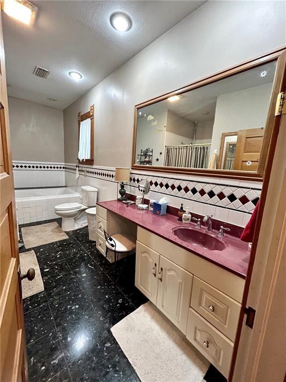 full bath with tile walls, vanity, tiled tub, toilet, and wainscoting