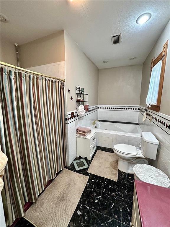 bathroom featuring tile walls, a bath, toilet, tile patterned flooring, and wainscoting
