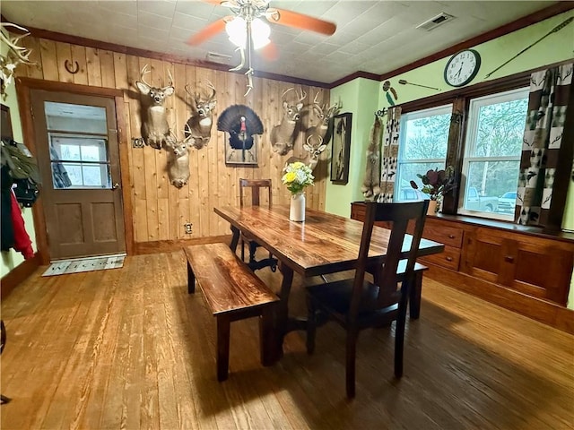 dining space with ceiling fan, wooden walls, crown molding, visible vents, and hardwood / wood-style floors
