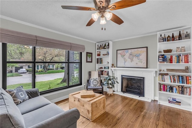living area with wood finished floors, ornamental molding, a brick fireplace, baseboards, and a ceiling fan