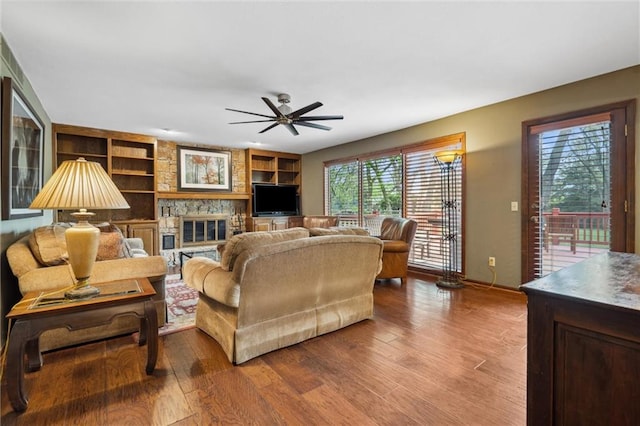 living room featuring a stone fireplace, built in features, ceiling fan, and wood finished floors